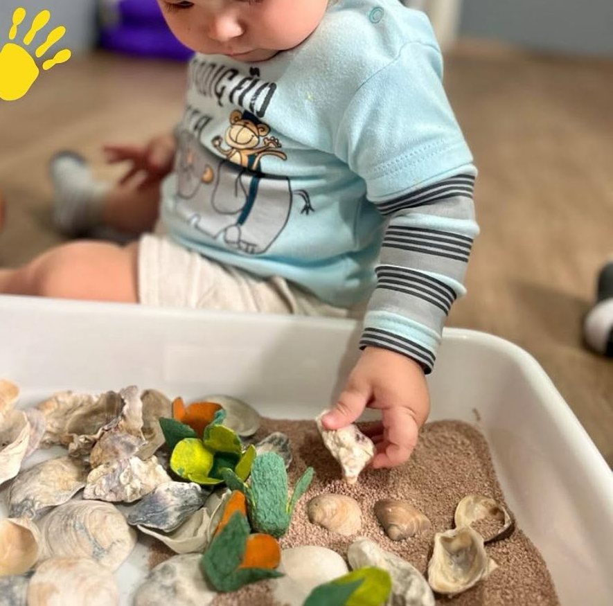 Bebê explorando caixa sensorial com areia e conchas no berçário